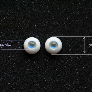 May include: Two white doll eyes with blue and green irises are displayed against a black, speckled background. The image includes labels for "Iris Size" and "Eye Size", indicating dimensions. The eyes are round and have a reflective quality.