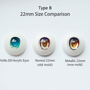 May include: Three 22mm doll eyes arranged horizontally. The left is labeled Volks DD Acrylic Eyes with a blue iris. The center is labeled Normal 22mm (old mold) with a brown iris. The right is labeled Metallic 22mm (new mold) with a gold iris.