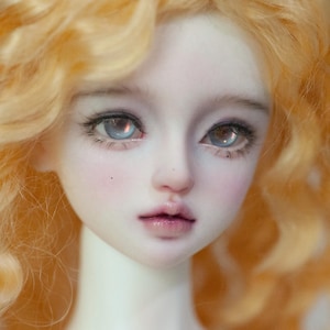 May include: Close-up of a doll's face with pale skin, rosy cheeks, and full lips. The doll has large, light blue eyes with detailed eyelashes and light orange, curly hair. The doll's expression is serene.