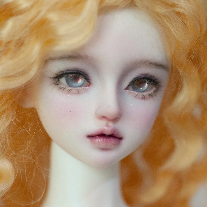 May include: Close-up of a doll's face with pale skin, rosy cheeks, and full lips. The doll has large, light blue eyes with detailed eyelashes and light orange, curly hair. The doll's expression is serene.