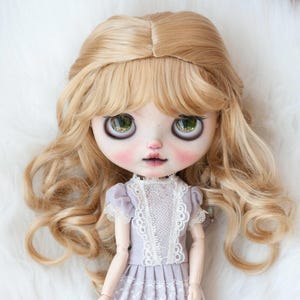 May include: A doll with long, wavy, blonde hair and bangs. The doll is wearing a light purple dress with white lace trim.