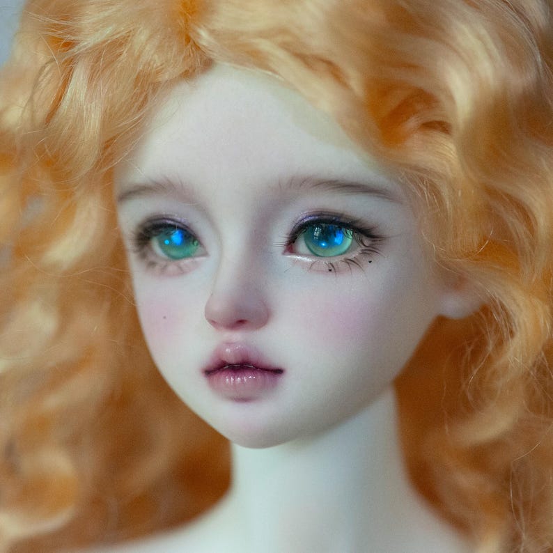 May include: A close-up of a doll's face with bright blue eyes and long eyelashes. The doll has pale skin, rosy cheeks, and light orange curly hair. The lips are a soft pink, and the overall aesthetic is delicate and detailed.