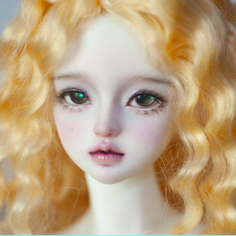 May include: A close-up of a doll's face with pale skin, rosy cheeks, and full lips. The doll has large, expressive eyes with long eyelashes and light brown irises. The doll's wavy, golden hair frames the face. The overall aesthetic is delicate and ethereal.