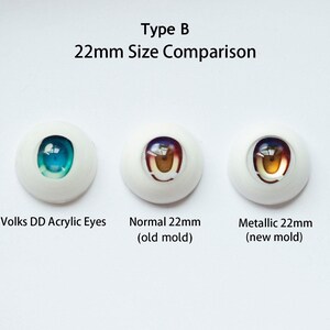 Puede incluir: Tres ojos de mu&ntilde;eca de 22mm dispuestos horizontalmente. El izquierdo etiquetado como Volks DD Acrylic Eyes con iris azul. El central como Normal 22mm (old mold) con iris marr&oacute;n. El derecho como Metallic 22mm (new mold) con iris dorado.