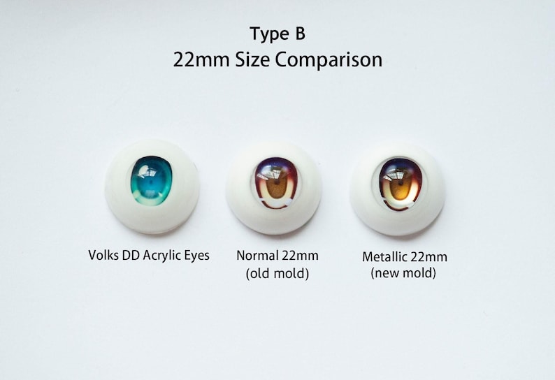 Puede incluir: Tres ojos de mu&ntilde;eca de 22mm dispuestos horizontalmente. El izquierdo etiquetado como Volks DD Acrylic Eyes con iris azul. El central como Normal 22mm (old mold) con iris marr&oacute;n. El derecho como Metallic 22mm (new mold) con iris dorado.