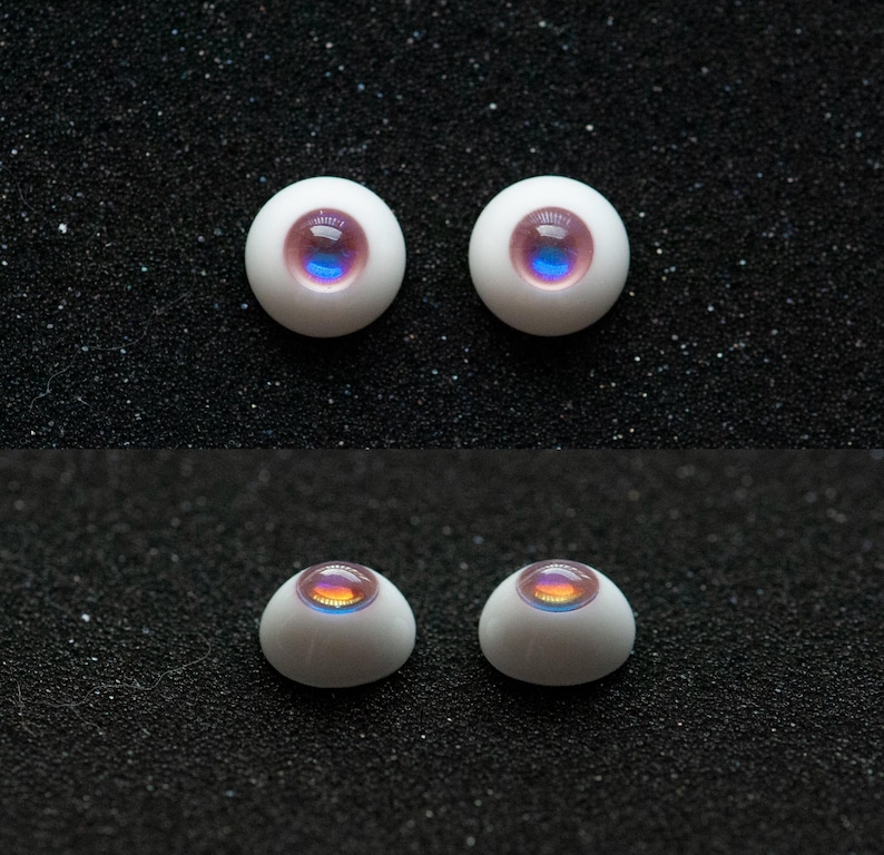 May include: Four white doll eyes with iridescent pink, blue, and yellow centers. The eyes are round and have a glossy finish. Two pairs of eyes are shown, each pair arranged side by side against a dark, textured background.