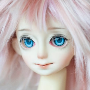 Puede incluir: Una cabeza de mu&ntilde;eca articulada con piel p&aacute;lida, grandes ojos azules vibrantes con pesta&ntilde;as detalladas y cabello rosa suave. Los rasgos faciales incluyen una nariz peque&ntilde;a, cejas definidas y labios brillantes, logrando una apariencia realista.