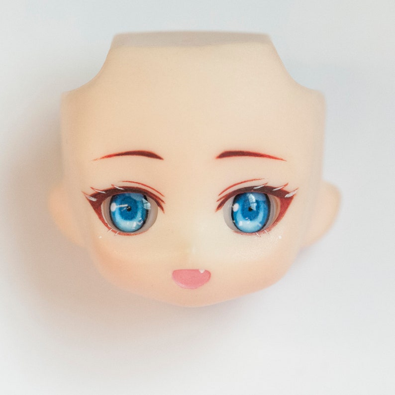 Puede incluir: Una cabeza de mu&ntilde;eca de tono de piel p&aacute;lido con ojos azules grandes y vibrantes con reflejos blancos detallados, cejas finas de color marr&oacute;n y una boca peque&ntilde;a abierta que muestra una lengua rosa. Los rasgos est&aacute;n pintados en mate.