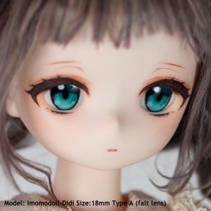 May include: Close-up of a doll's face with blue eyes and a light pink complexion. The doll has brown hair and is wearing a white dress. Model: Imomodol-Didi Size: 18mm Type A (falt lens)