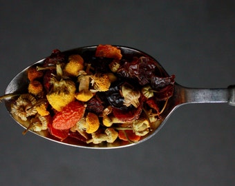 May include: A silver spoon filled with a blend of dried herbs and flowers, including red goji berries, yellow chamomile flowers, and dark red hibiscus petals.