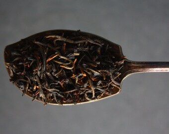 May include: A silver spoon filled with loose leaf black tea. The tea leaves are a mix of dark brown and black.