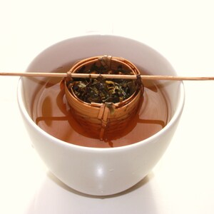 May include: A white ceramic teacup with a brown bamboo tea infuser filled with loose leaf tea. The infuser is suspended in the teacup by a wooden stick.
