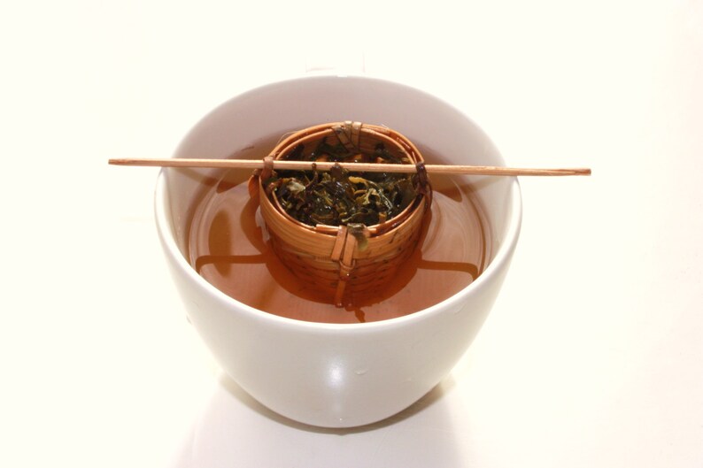 May include: A white ceramic teacup with a brown bamboo tea infuser filled with loose leaf tea. The infuser is suspended in the teacup by a wooden stick.