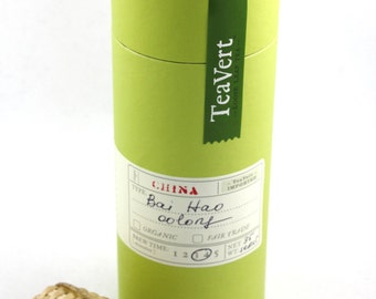May include: A green cylindrical tube with a label that reads "TeaVert Loose Leaf Tea" and "Bai Hao oolong" in handwritten text. The tube is sitting on a white surface next to a small bamboo tea infuser.