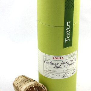 May include: A green cylindrical container of TeaVert loose leaf tea, labeled "India Poobong Darjeeling 1st Flush". The container has a white label with checkboxes for "Organic" and "Fair Trade". The container is sitting on a white surface next to a bamboo tea infuser.