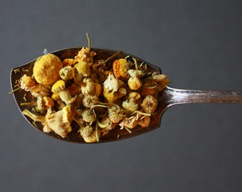 May include: A silver spoon filled with dried chamomile flowers. The flowers are a mix of yellow and white.