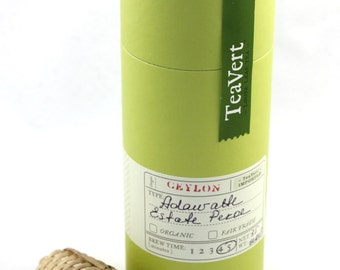 May include: A green cylindrical tea tin with a white label that reads "CEYLON Adawash Estate Pekoe" and "Tea Vert Loose Leaf Tea". The label also includes a check box for "Organic" and "Fair Trade". The tin has a bamboo tea infuser with a wooden handle.