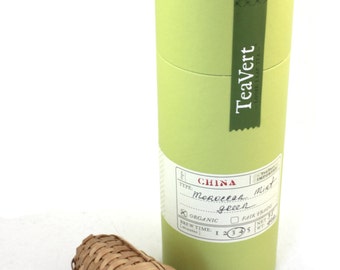 May include: A green cylindrical tube with a label that reads "TeaVert Loose Leaf Tea" and "China Moroccon Mint Green" with a checkmark next to "Organic" and a checkmark next to "Fair Trade". The tube is sitting on a white surface next to a brown woven tea infuser.