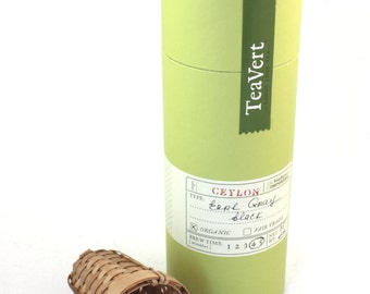 May include: A light green cylindrical container of "TeaVert" loose leaf tea with a label indicating "Ceylon Earl Gray Black" tea. A small woven tea strainer rests in front of the container.