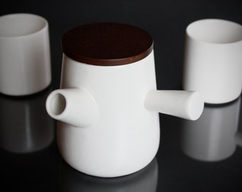 May include: White ceramic teapot with a wooden lid and two handles. The teapot is sitting on a black surface.