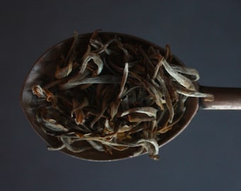 May include: A close-up of a spoon filled with loose leaf white tea. The tea leaves are a light brown color and have a slightly dried appearance.