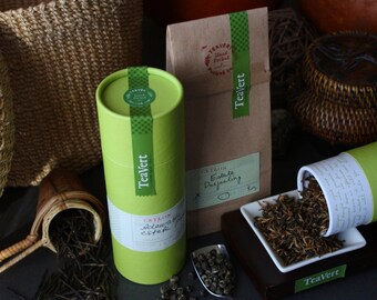 May include: Two green tea canisters with the text "TeaVert" and "Crayola Estate Darjeeling" on them. The canisters are sitting on a brown surface with a white plate of loose leaf tea.