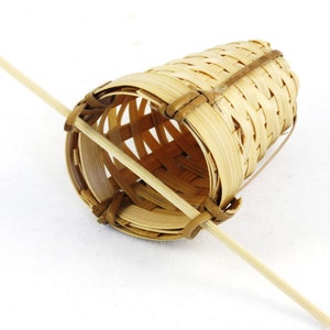 May include: A woven bamboo tea infuser with a wooden handle. The infuser is cylindrical and has a woven pattern. The handle is attached to the infuser at the top and bottom.