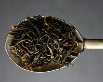 May include: A silver spoon filled with loose leaf black tea. The tea leaves are a mix of dark brown and golden yellow.