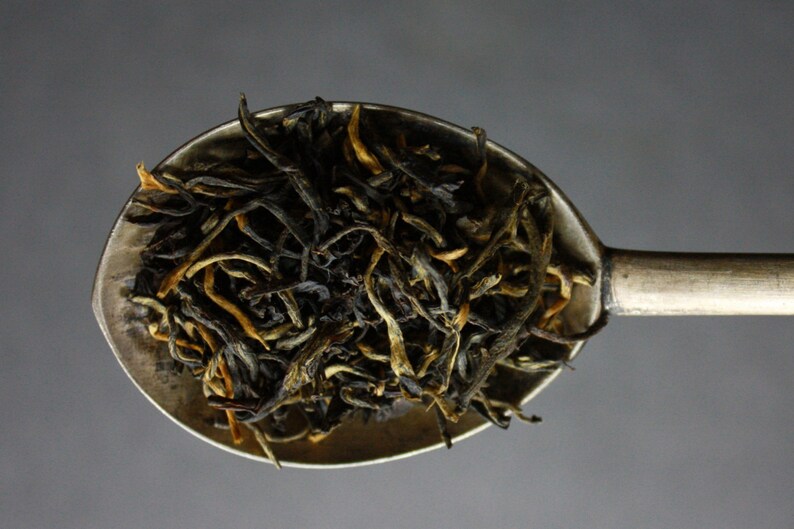 May include: A silver spoon filled with loose leaf black tea. The tea leaves are a mix of dark brown and golden yellow.