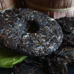May include: A dark brown, round tea cake with a hole in the center. The cake is made of compressed tea leaves and has a rough, textured surface. The cake is surrounded by loose tea leaves.