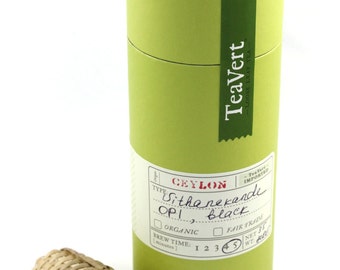 May include: A green cylindrical box with a white label that reads "TeaVert Loose Leaf Tea" and "CEYLON Sithanekande OPI, black". The label also includes checkboxes for "Organic" and "Fair Trade" and a brew time guide. The box is sitting on a white surface next to a bamboo tea infuser.