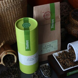 May include: Two green tea canisters with the text "TeaVert" and "Crayola Estate Darjeeling" on them. The canisters are sitting on a brown surface with a white plate of loose leaf tea.