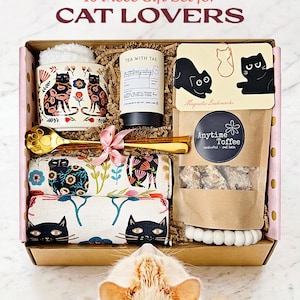 May include: A 10-piece gift set in a cardboard box featuring a mug with cat illustrations, a Tea With Tae canister, a gold paw-print spoon, two patterned cloths with cat motifs, a bag of Anytime Toffee, and magnetic bookmarks with cat designs.