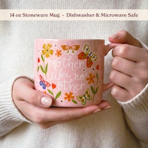 A person is holding a pink mug with a floral design and butterflies, and the text "mother like no other" is printed on it.