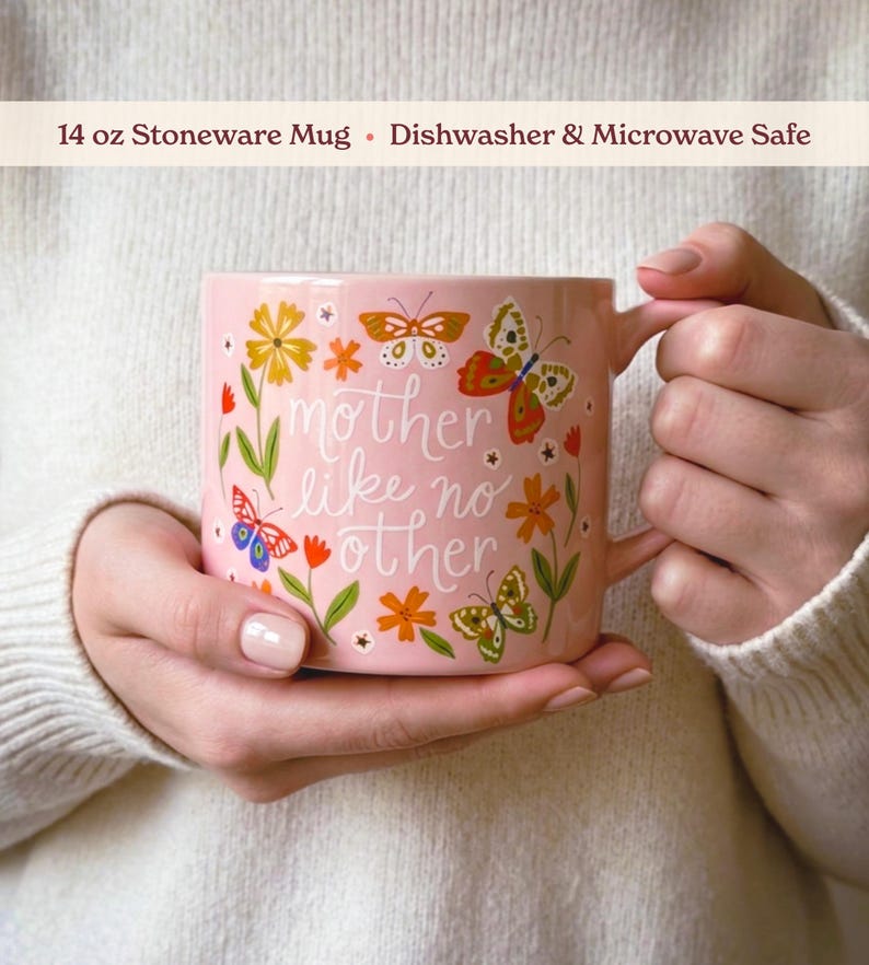 A person is holding a pink mug with a floral design and butterflies, and the text "mother like no other" is printed on it.