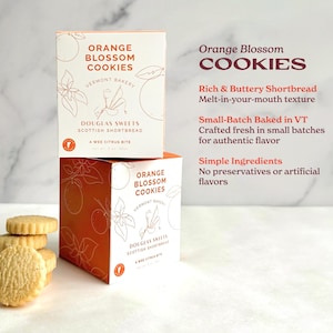 a box of Vermont Bakery's Orange Blossom Cookies, with a stack of cookies placed next to it. The box is orange and white, and the cookies are white.