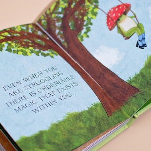 An open page from the illustrated book showing a whimsical frog swinging from a tree branch. The text reads: 'Even when you are struggling there is undeniable magic that exists within you.'