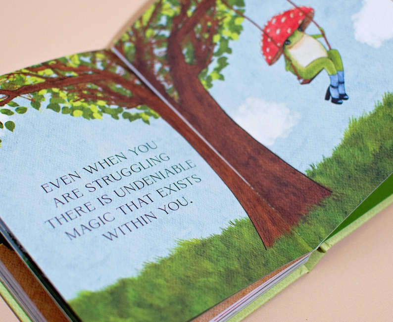 An open page from the illustrated book showing a whimsical frog swinging from a tree branch. The text reads: 'Even when you are struggling there is undeniable magic that exists within you.'