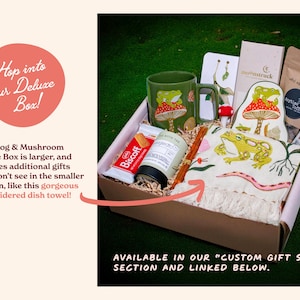 Expanded deluxe version of the frog and mushroom themed gift set displayed on a forest green background. Includes an embroidered dish towel alongside the mug trinket tray jasmine green tea and snacks. Designed for thoughtful gifting and relaxing woodland-inspired afternoons.