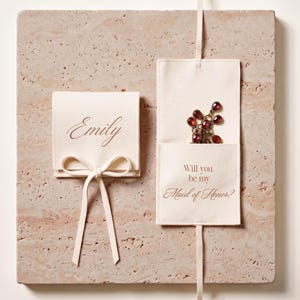 May include: Two off-white fabric pouches with text and a bow, set on a textured stone surface. One pouch says "Emily" in script. The other says "Will you be my Maid of Honor?" and holds a red gemstone necklace.