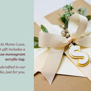 Brown kraft paper gift box wrapped in an elegant cream satin ribbon with a sprig of dried greenery. Attached to the bow is a personalized custom gold acrylic monogram initial tag shaped like the letter S.