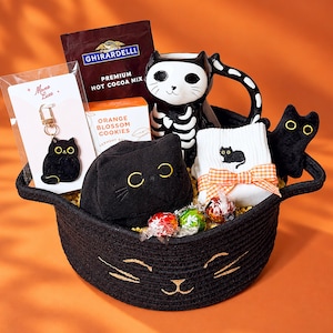 May include: A black woven basket with cat face embroidery containing a Ghirardelli hot cocoa mix, an orange blossom cookies box, a skeleton cat mug, a black cat keychain, a black plush cat, white socks with a cat, and three wrapped candies.