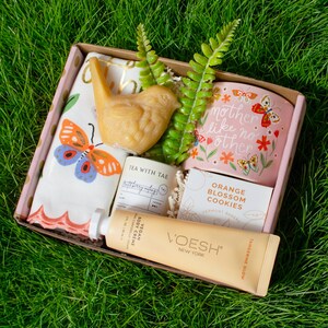 A gift box containing various items, including a small bird figurine, a tea kettle, a cookie, and a tube of cream, all placed on a grassy surface.