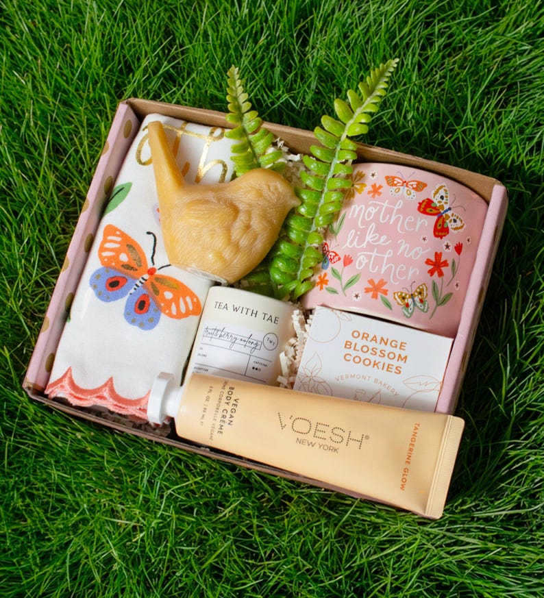A gift box containing various items, including a small bird figurine, a tea kettle, a cookie, and a tube of cream, all placed on a grassy surface.
