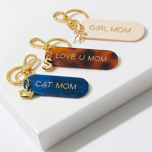 May include: Three keychains with gold-colored hardware. One is light pink with "GIRL MOM" text, another is tortoiseshell with "LOVE U MOM", and the last is blue with "CAT MOM". Each keychain has a charm.