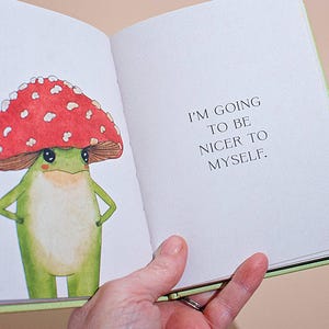 An open page from the self-care book showing a colorful watercolor illustration of a frog wearing a red mushroom cap next to the text, 'I'm going to be nice to myself.'