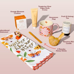 A collection of gifts including an embroidered dishtowel with the text "mother like no other", a pink stoneware mug, orange blossom cookies, and a beeswax bird candle. Also included are a tube of vegan body creme, fruit & oolong tea, and honey sticks.