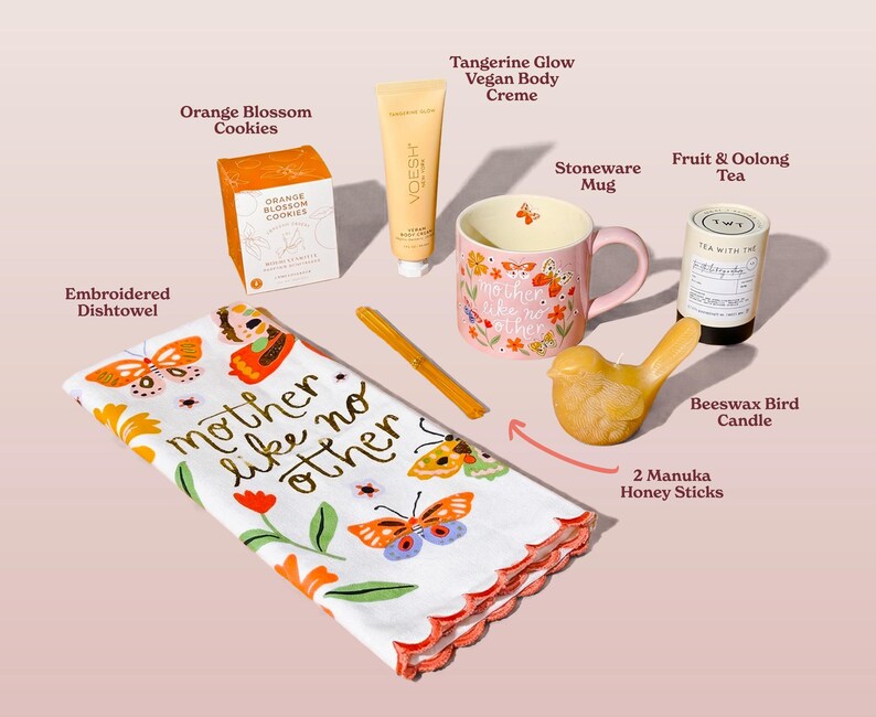 A collection of gifts including an embroidered dishtowel with the text "mother like no other", a pink stoneware mug, orange blossom cookies, and a beeswax bird candle. Also included are a tube of vegan body creme, fruit & oolong tea, and honey sticks.