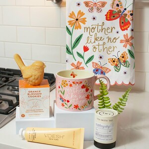 a kitchen counter with various items, including a bird figurine, a pink mug with a floral design, a tea kettle, a tube of Voesh New York, and a decorative towel with a butterfly design.