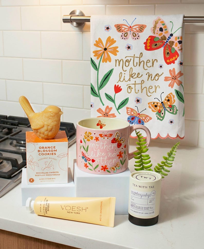 a kitchen counter with various items, including a bird figurine, a pink mug with a floral design, a tea kettle, a tube of Voesh New York, and a decorative towel with a butterfly design.